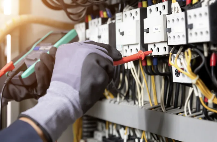 A person wearing protective gloves uses a screwdriver to adjust wiring in an electrical panel filled with cables and circuit breakers at a site prepared by a demolition company in New York NY.
