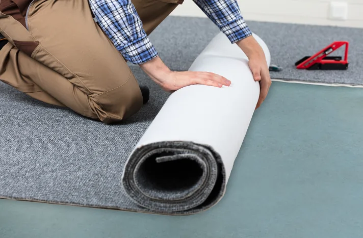A person kneels on the floor while unrolling a gray carpet onto a blue surface, possibly as part of a demolition in NYC. A utility knife and a red measuring tool are visible in the background.