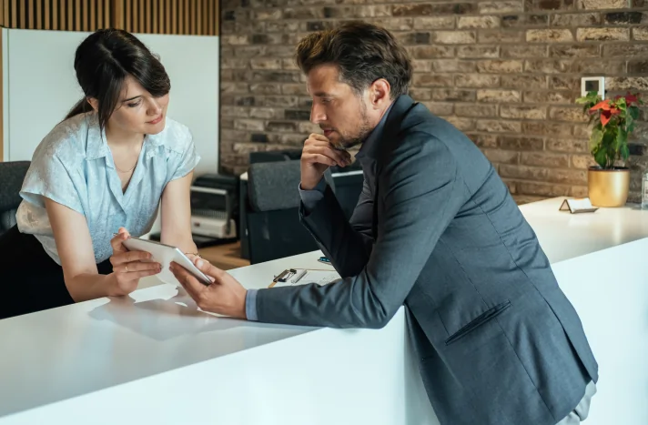 A woman and a man stand at a modern reception desk, both looking at a digital tablet as they discuss demolition services in NYC. The man appears thoughtful, surrounded by office equipment and a potted plant.
