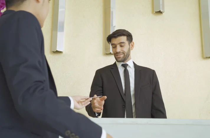A receptionist in a suit stands behind a counter, smiling as he hands a key card to a guest who is also wearing a suit, while a brochure about demolition services in NYC sits neatly on the counter.