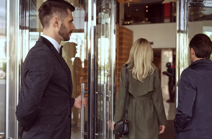 A man in a suit holds a glass door open as a blonde woman in a green coat and a man in a dark jacket, perhaps discussing demolition services in NYC, walk into a modern building with wood and glass decor.
