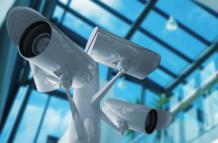 Close-up of three white security cameras mounted on a single pole, angled in different directions, inside a modern building—ideal for monitoring safety during demolition services in NYC. Large glass windows fill the space with bright natural light.