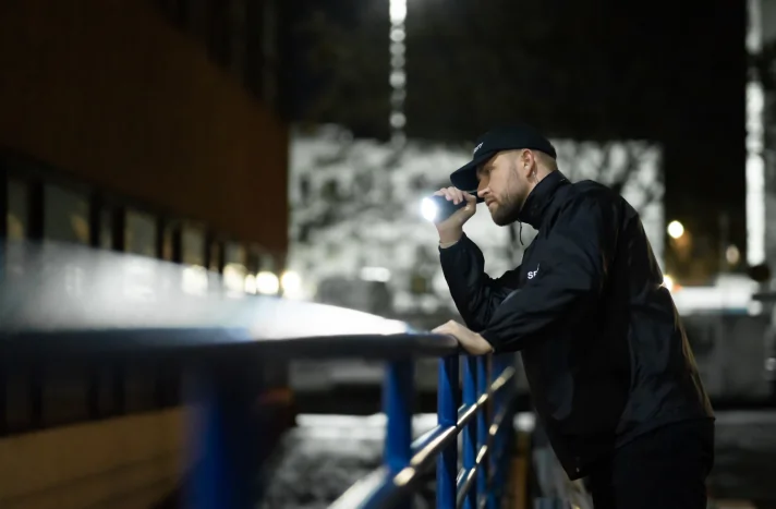 A security guard in a black uniform and cap leans on a blue railing at night, holding a flashlight and surveying the site of a recent demolition in Brooklyn NY, with blurred city lights in the background.