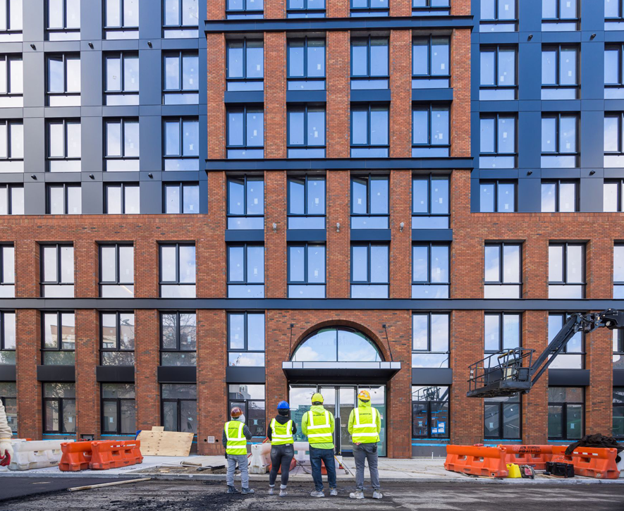 Four construction workers in safety vests and helmets stand facing a modern brick and glass building under construction, with orange barricades and equipment from a demolition company in New York NY visible in the foreground.