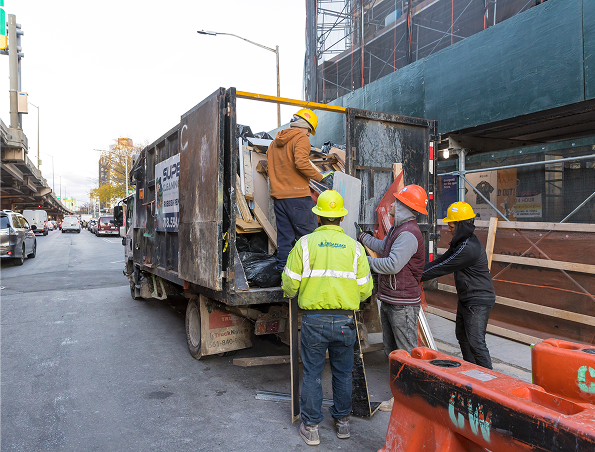Four construction workers in safety gear load debris and materials into a dump truck on a city street lined with buildings and scaffolding. Traffic and pedestrians are visible in the background.