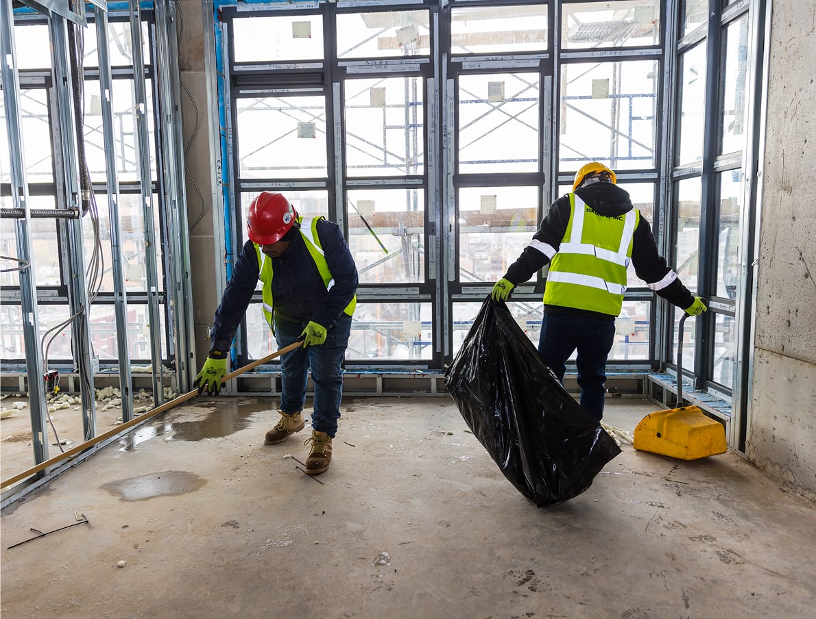 Two construction workers wearing safety vests and helmets clean inside a building under construction; one sweeps debris while the other holds a large black trash bag and a dustpan, with scaffolding visible through windows.