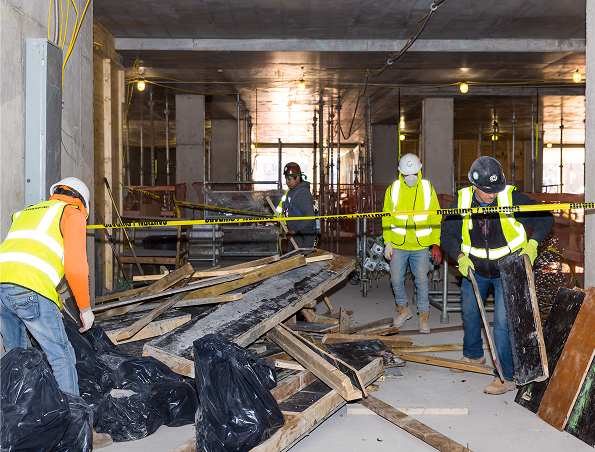Four construction workers in safety vests and helmets clean up wooden debris and trash bags inside a building under construction, with caution tape and exposed wiring visible.