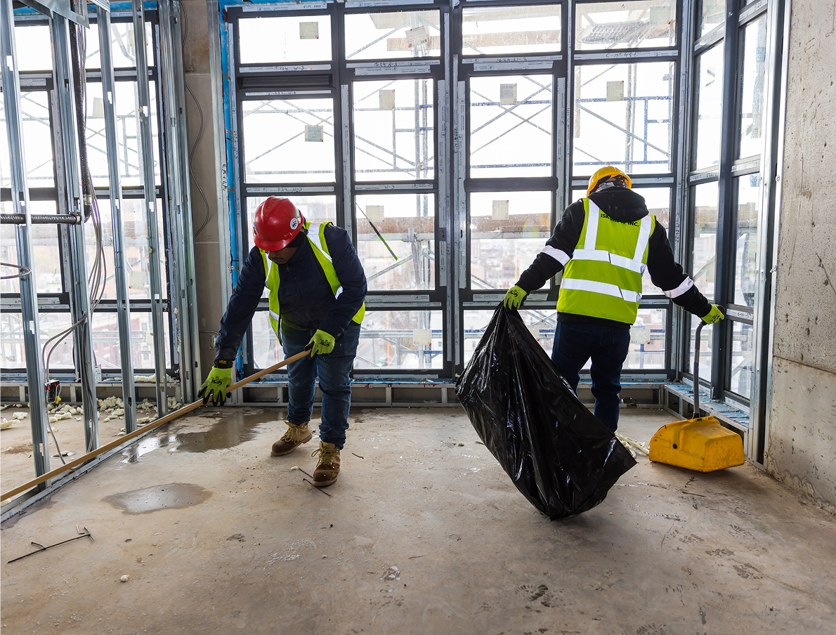 Two construction workers in safety vests and helmets clean a building site. One sweeps debris into a dustpan while the other holds a large black trash bag. Large windows and scaffolding are visible in the background.