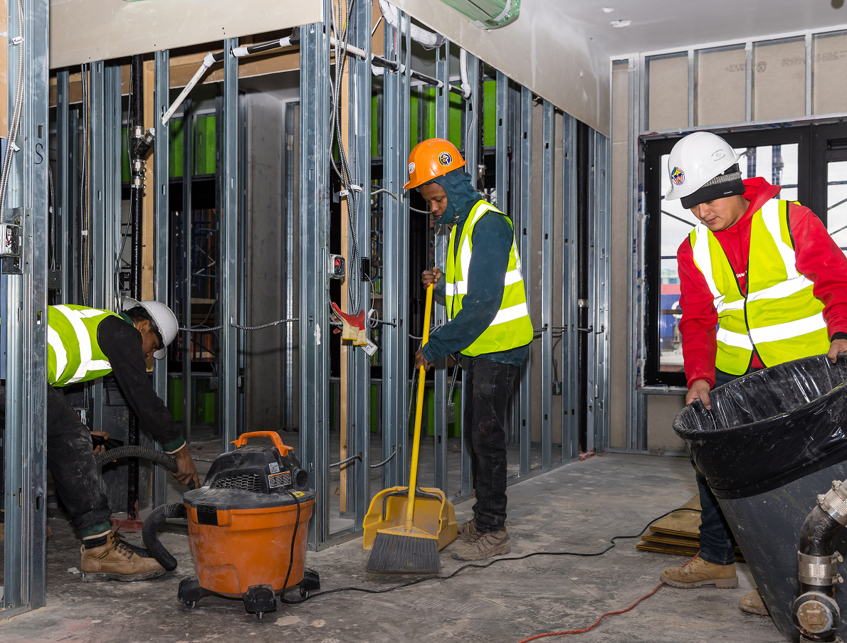 Three construction workers wearing safety vests and helmets clean a building under construction. One uses a vacuum, another sweeps with a broom and dustpan, and the third holds a large black bucket. Exposed wall frames are visible.