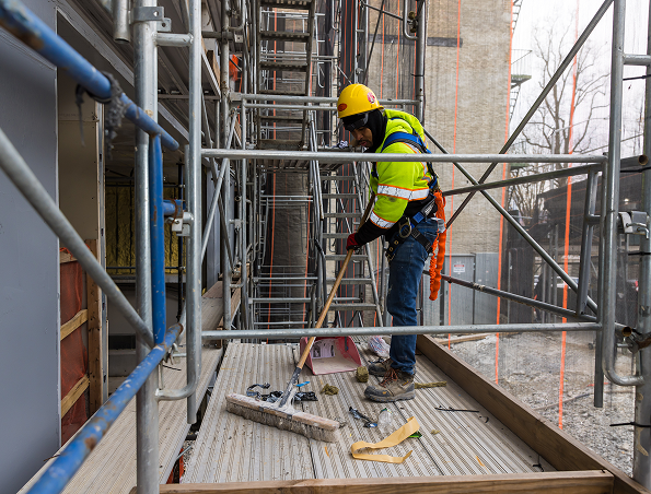A construction worker in safety gear and a helmet stands on scaffolding, using a broom to sweep debris and scattered tools on a wooden platform at a building site.