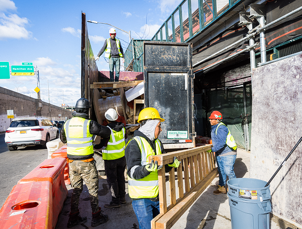 Five construction workers wearing safety vests and helmets move a wooden barrier and work near a building site beside a road. One worker stands above them on a ramp while others lift and carry materials.