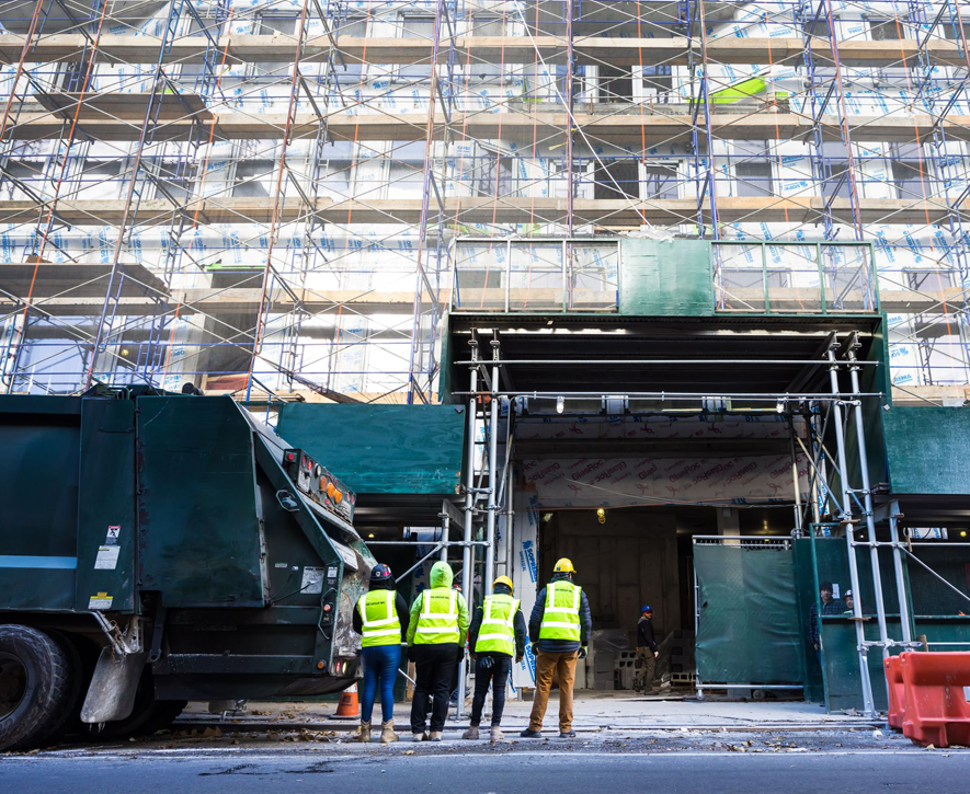 Four construction workers in safety vests and helmets stand in front of a building under construction, with scaffolding covering the facade and a large truck parked nearby.
