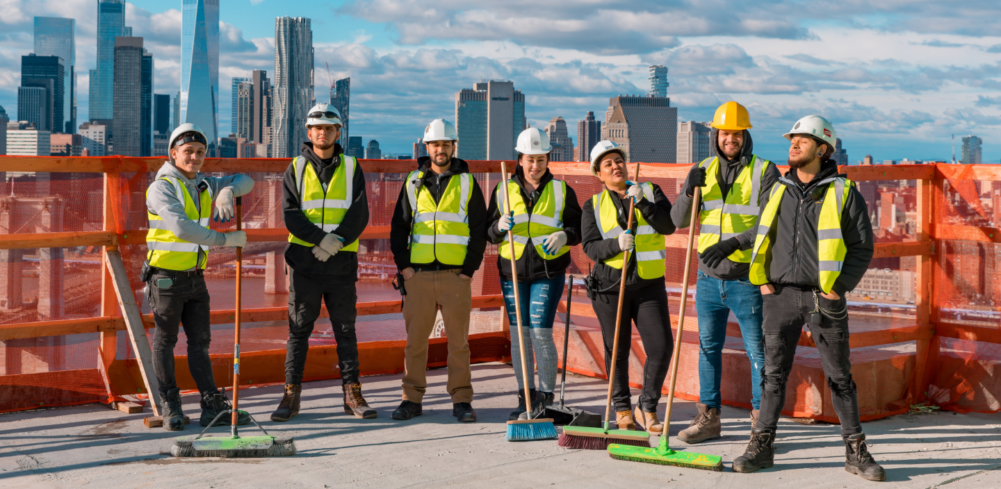 Six construction workers wearing safety vests and helmets stand on a rooftop with brooms. A city skyline with tall buildings is visible in the background under a partly cloudy sky.