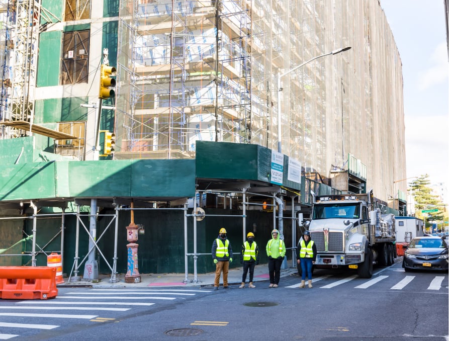 Four construction workers wearing safety gear stand at a crosswalk in front of a large building covered in scaffolding and netting, with a truck and taxi nearby on a sunny day.