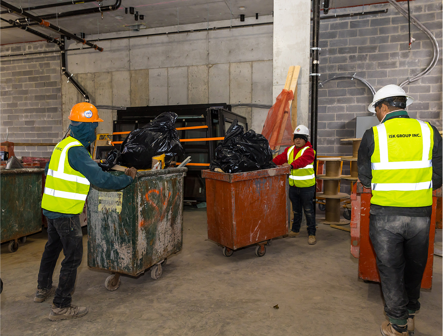 Three construction workers in safety gear and reflective vests move large bins filled with black trash bags inside an unfinished building with concrete walls and exposed pipes.