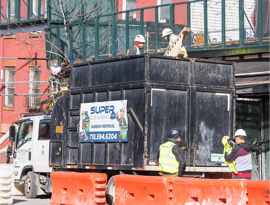 Four construction workers load debris into a large black rubbish removal container on a truck. Orange barriers and a &ldquo;Super Cleaning&rdquo; sign with contact details are visible. A red building and scaffolding are in the background.