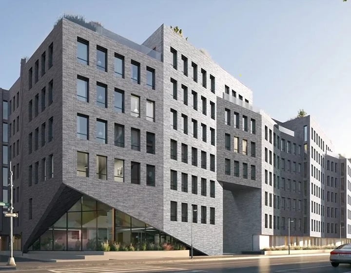 Modern apartment building with a geometric, angular design and gray brick exterior. Large windows line the facade, and a sloped entrance is visible at street level under a sunny sky.