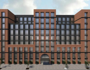 A modern, multi-story building with a symmetrical facade, large rectangular windows, and red brick exterior. The entrance has an arched doorway, and there are a few small trees along the sidewalk in front.