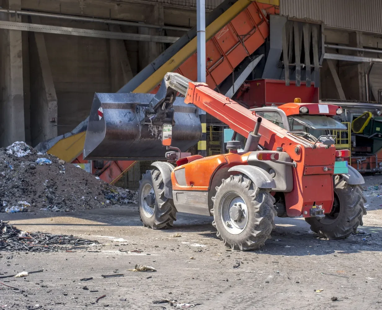 An orange telescopic handler with a front bucket is parked at an industrial site near piles of debris and a conveyor belt system, likely for waste processing or recycling.