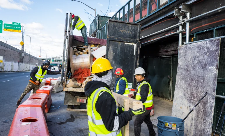Construction workers in safety vests and helmets unload materials from a truck near an overpass, surrounded by orange barriers and equipment from a leading demolition company in New York NY.