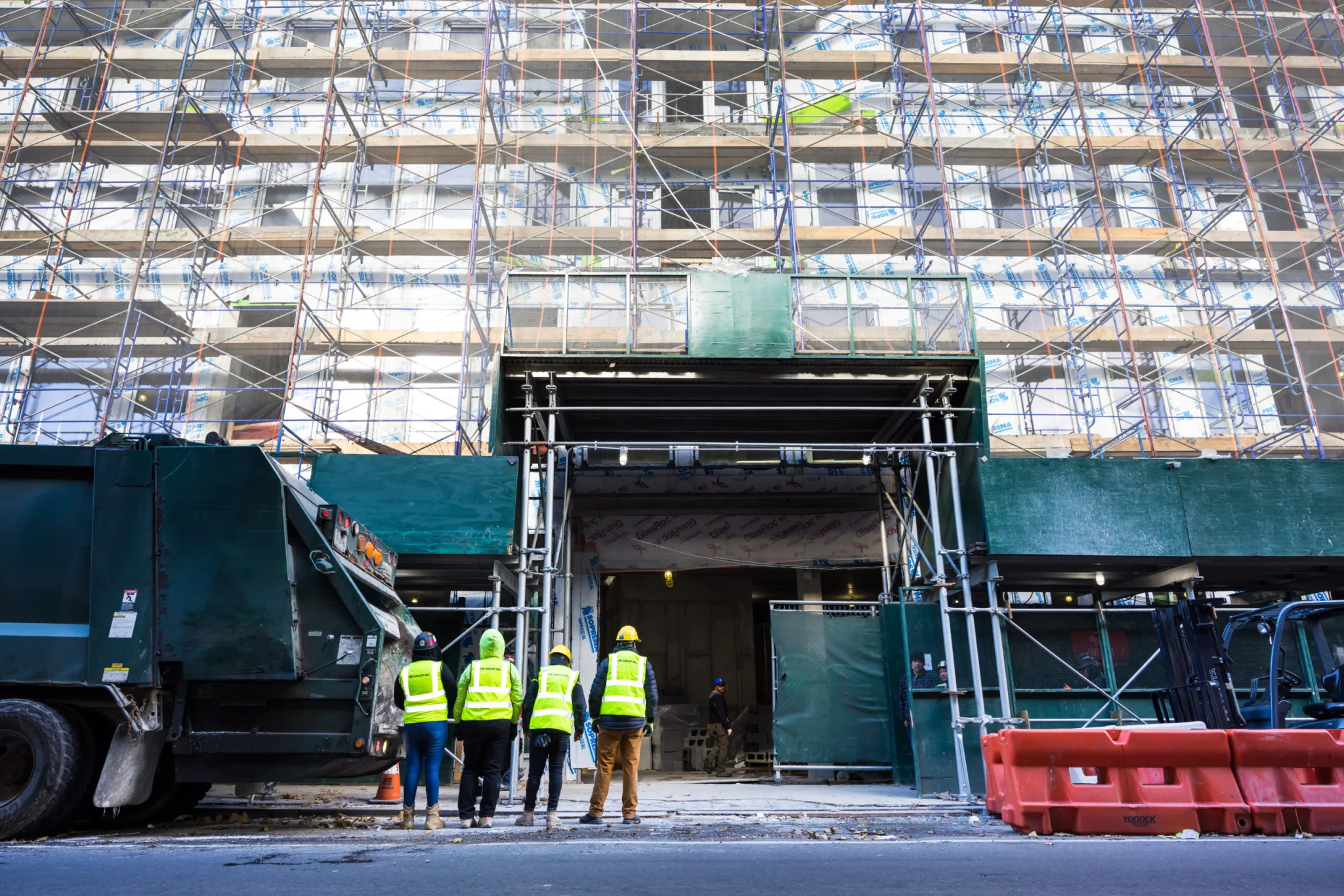 Construction workers in yellow vests and hard hats stand near a building covered in scaffolding, as a demolition company in New York NY operates vehicles and barriers along the street in front.