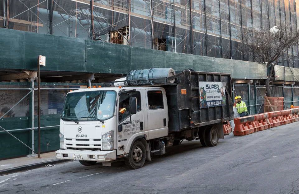 A white Isuzu dump truck with a black dumpster is parked on a city street beside a construction site. Two construction workers in safety gear stand near orange barriers and green scaffoldings, typical of demolition services in NYC.