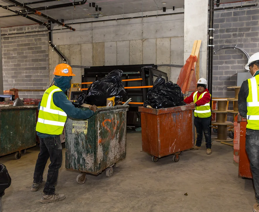 Two construction workers in safety vests and helmets move large bins filled with black garbage bags inside a building under construction, exemplifying the efficiency of demolition services in NYC amid exposed concrete walls and tools.