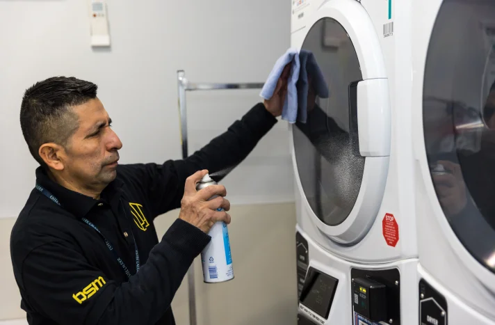 A man in a black uniform cleans the front of a washing machine with a cloth and cleaning spray inside a laundry room, keeping the space spotless after recent demolition in Manhattan NY.