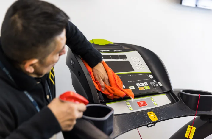 A person cleans the control panel of a treadmill with an orange cloth while holding a spray bottle, ensuring the machine is sanitized, much like a demolition company in New York NY ensures spaces are thoroughly cleared and prepared.