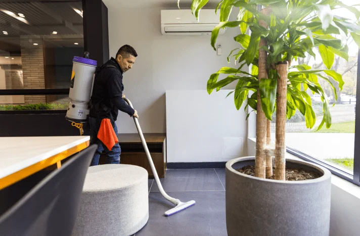 A man wearing a backpack vacuum cleans the floor in a bright, modern room with large windows and potted plants, restoring freshness after recent demolition in Manhattan NY.