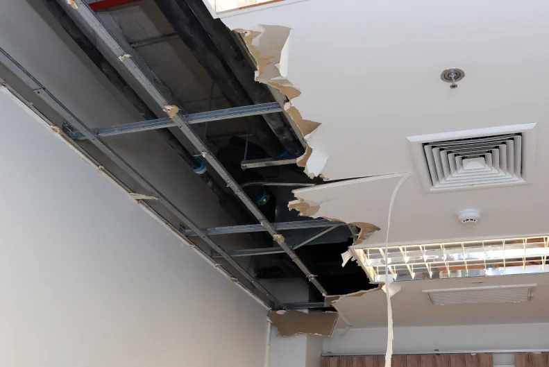 A ceiling with severe damage and missing panels exposes metal framework, ductwork, and wires above—signs often seen before demolition services in NYC begin. Pieces of broken ceiling hang down, and light fixtures are attached to the intact section.