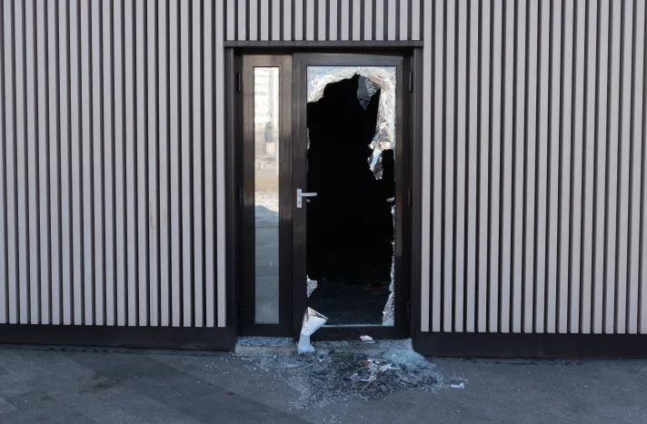 A glass door with a large, jagged hole and shattered glass around it is set in a building with vertical beige and brown stripes—damage often seen by any demolition company in New York NY. The interior beyond the door appears dark and empty.