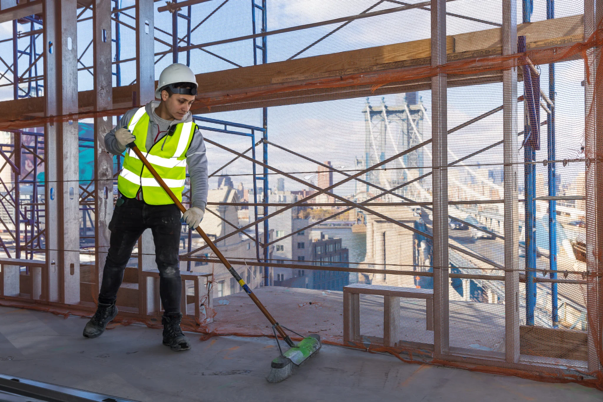 A construction worker wearing a hard hat and reflective vest sweeps the floor of an unfinished building. Scaffoldings and mesh surround the area, typical of sites managed by a demolition company in New York NY, with a large bridge visible in the background.