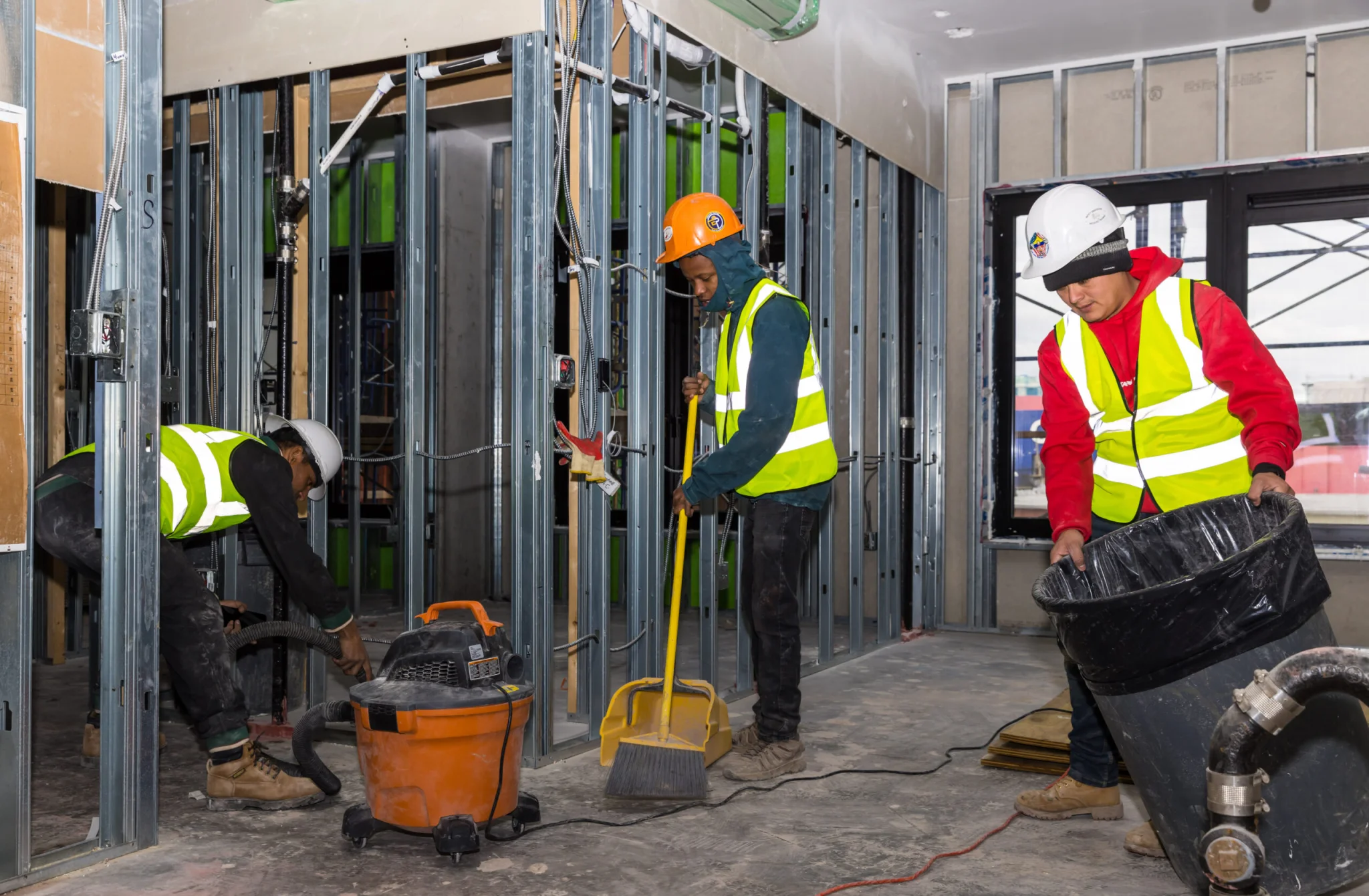 Three construction workers in safety vests and hard hats clean a building site after demolition services in NYC. One vacuums the floor, another sweeps with a broom and dustpan, and the third carries a large trash bin. Exposed metal framing is visible.